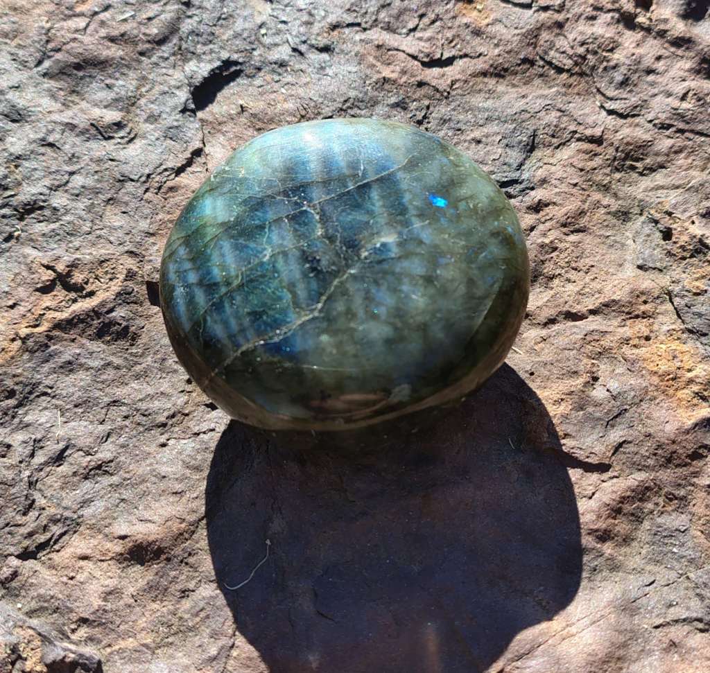Labradorite palm stone with blue-green flashes and natural grey tones resting on a rock outdoors.